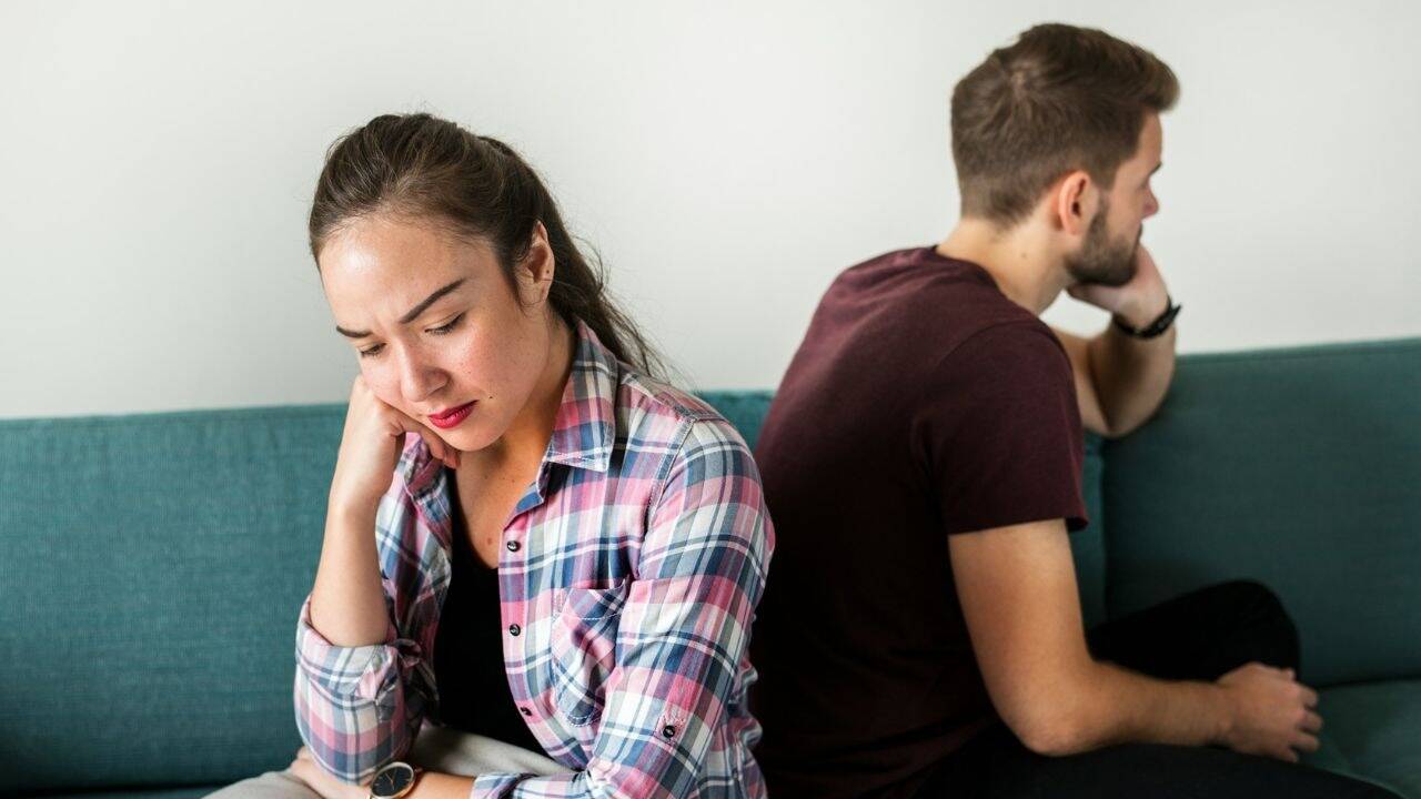 A man and a woman sit on a couch with their backs to each other. The woman looks down, while the man looks away.