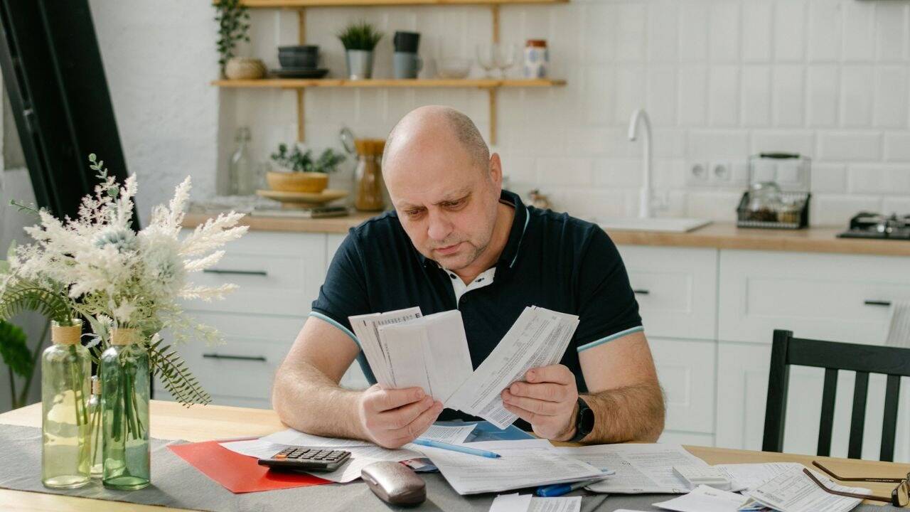 A serious bald man sits at a table, looking at papers and a calculator.