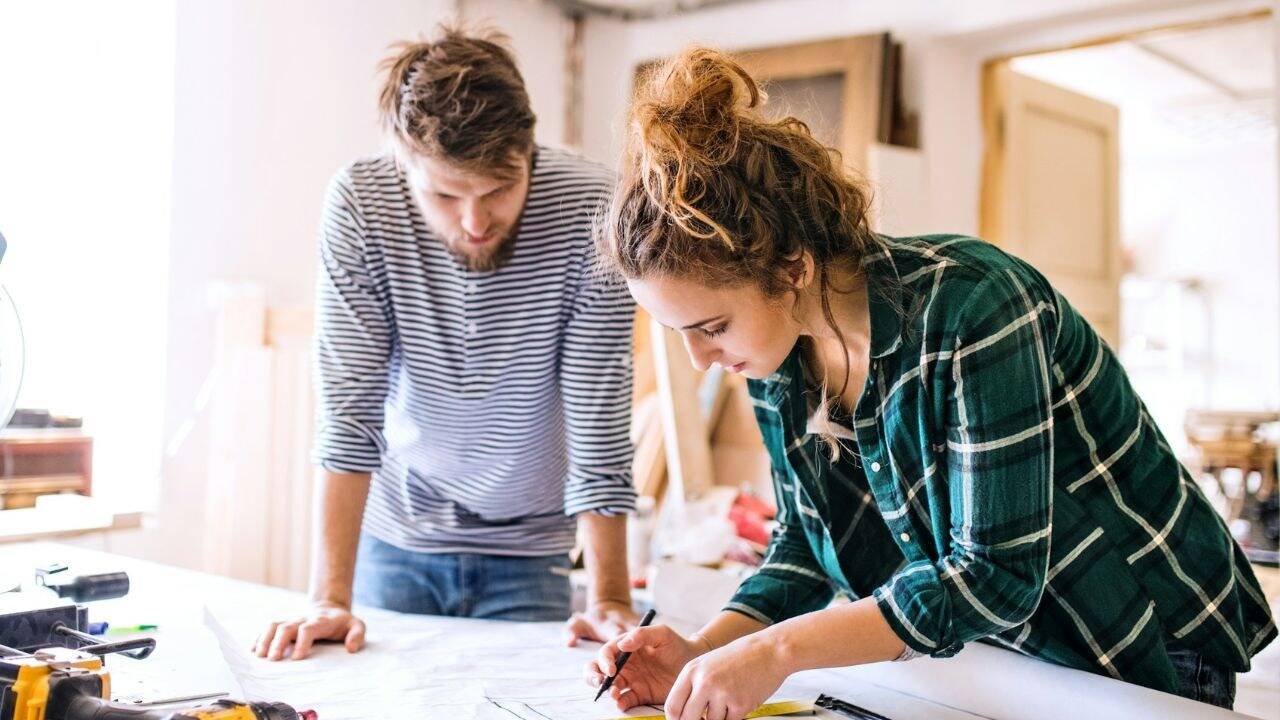 A couple stands over a large sheet of paper, with the woman holding a pencil and a tape measure.
