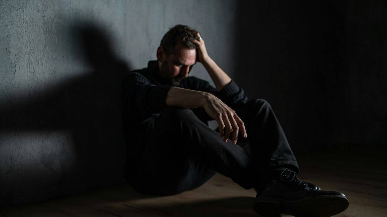A stressed man sits against a wall, holding his head with his hand and looking down.