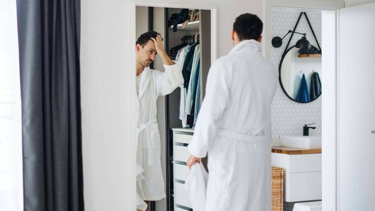 A man in a white robe looks in a mirror, adjusting his hair in a closet.