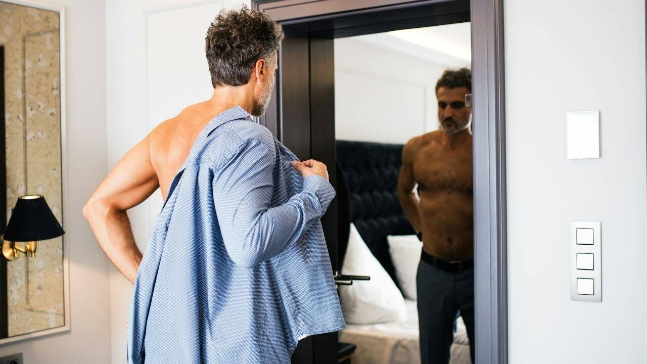 A shirtless man looks in a mirror, putting on a light blue button-down shirt.