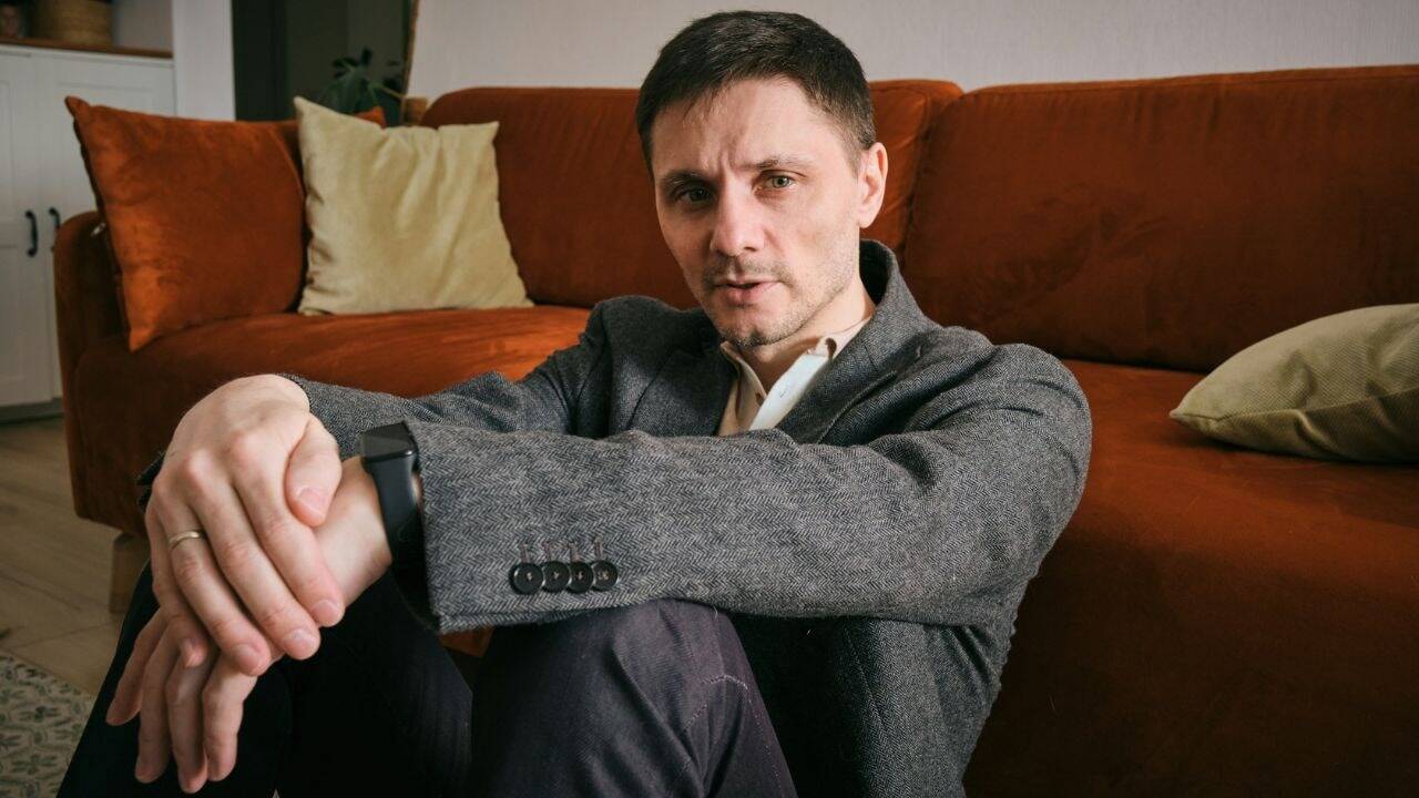 A man in a gray jacket and a watch sits on the floor, looking at the camera.