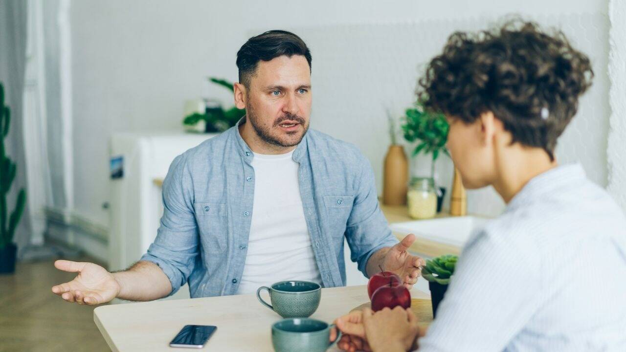 A man sits at a table, gesturing with his hands and talking to another person.