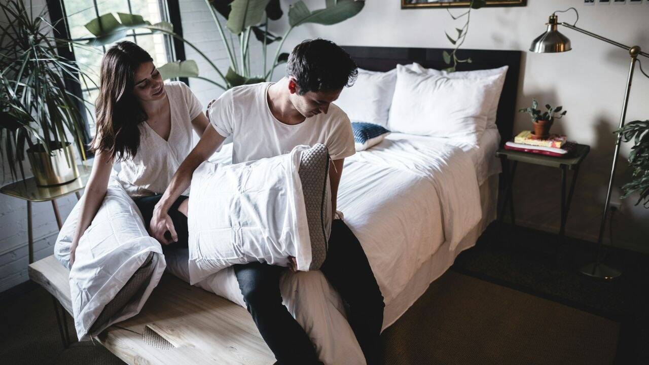 A couple sits on a wooden bench, holding pillows and looking at each other.