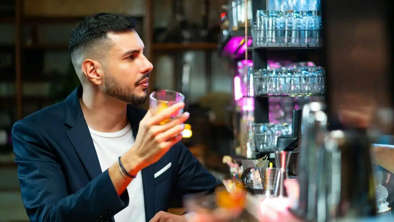 A well-dressed man holding a glass sits alone at a brightly lit bar counter.