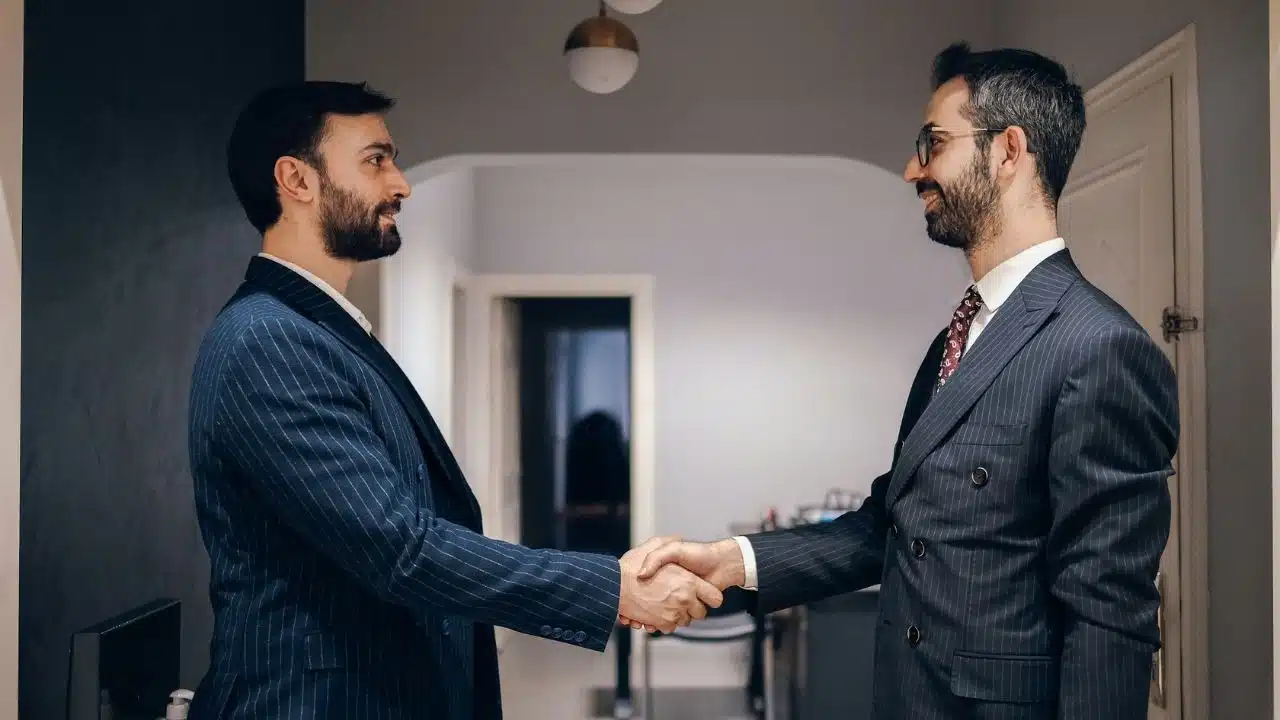 Two formally dressed men with beards shaking hands in an indoor hallway.