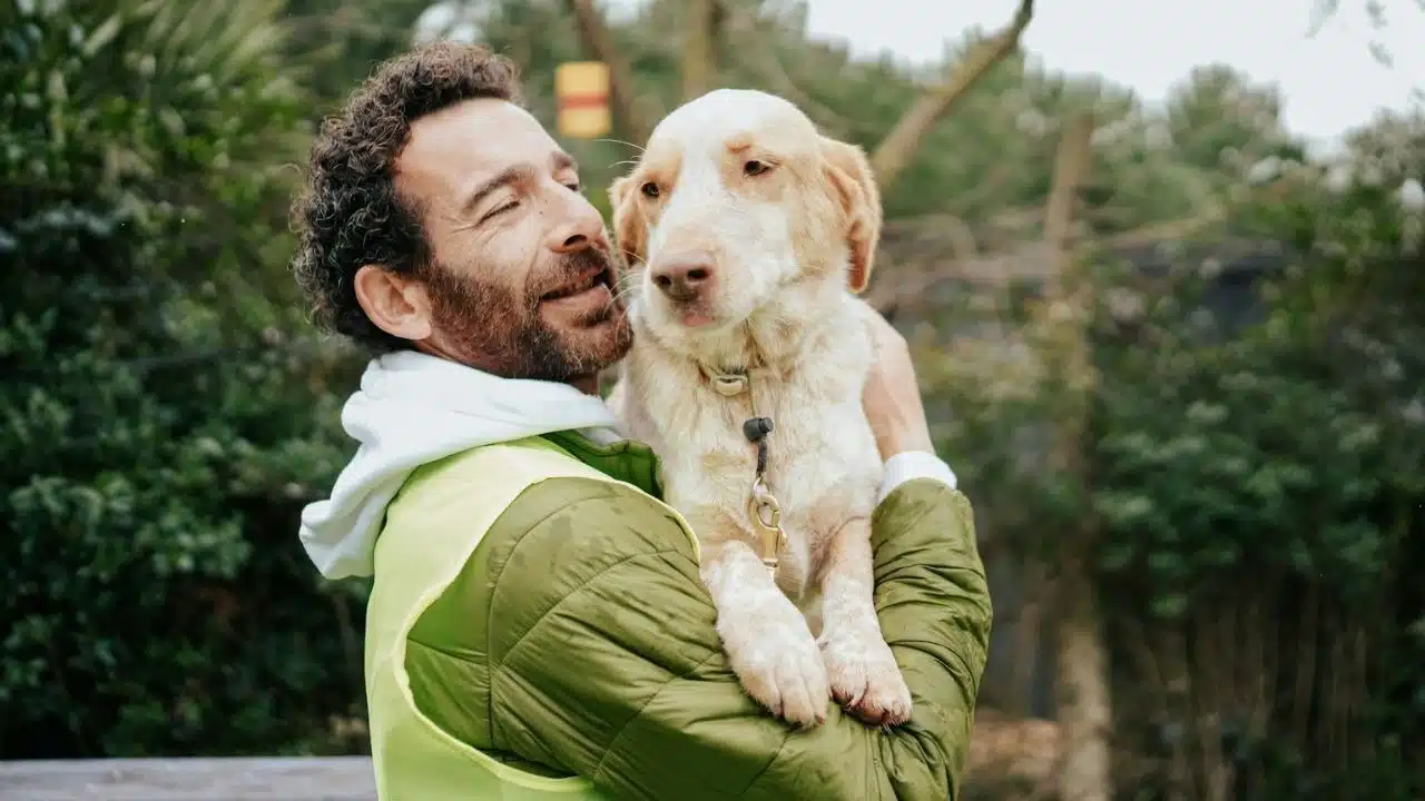 Man in a green jacket smiles while holding a light-colored dog outdoors.