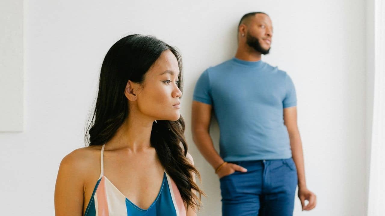 Woman in the foreground looks left while a blurry man leans against a wall in the background.