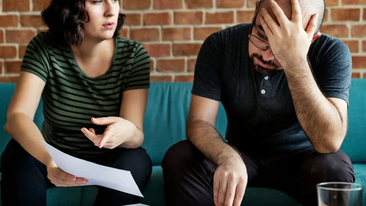 A man sits with his head in his hand, looking stressed, while a woman holding papers talks to him.