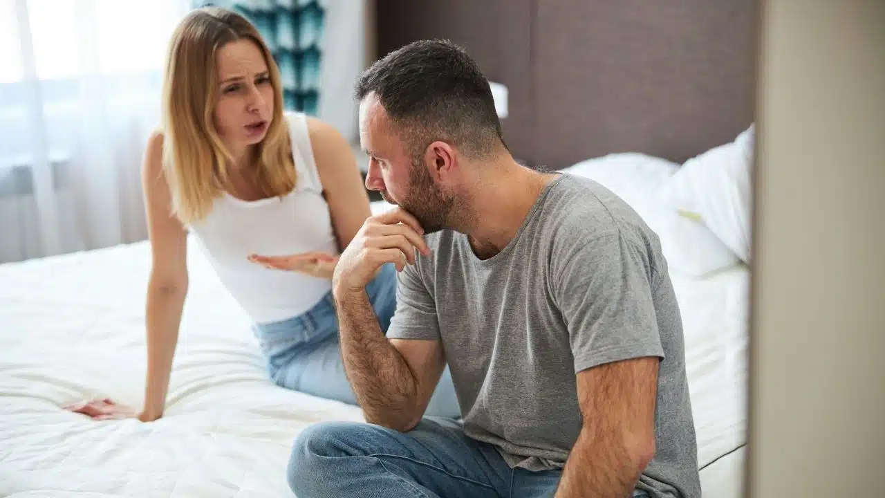 A man in a gray t-shirt sits with his head down while a woman in a white tank top gestures and speaks in the background.