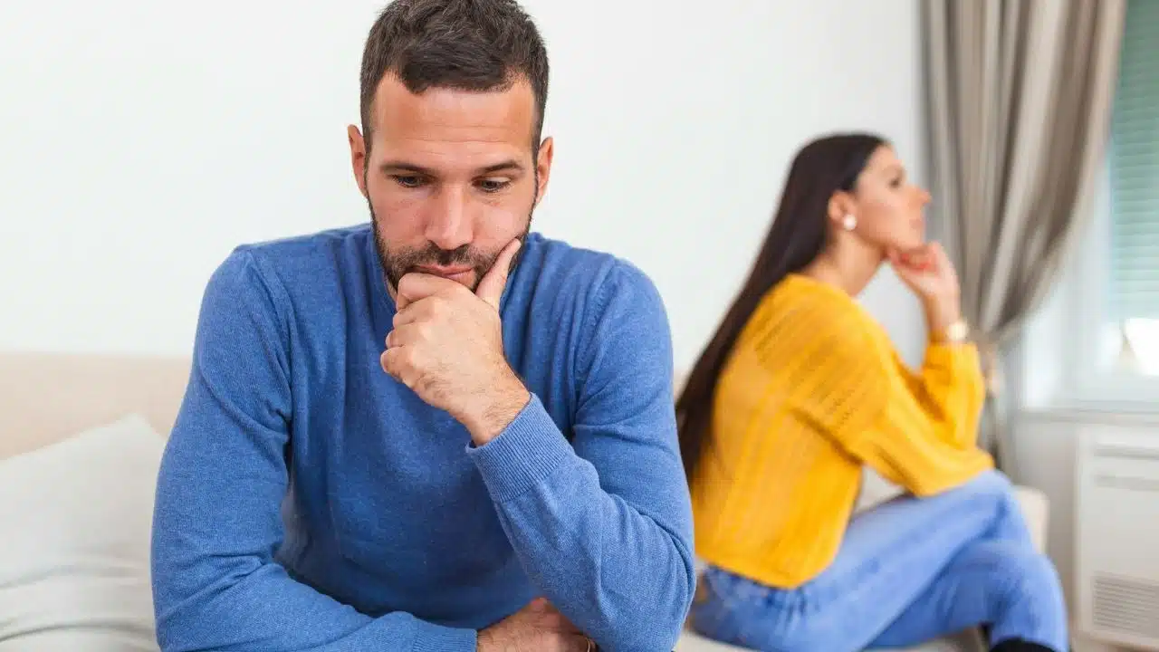 A man in a blue sweater sits in the foreground, looking sad, while a woman in yellow sits behind him.