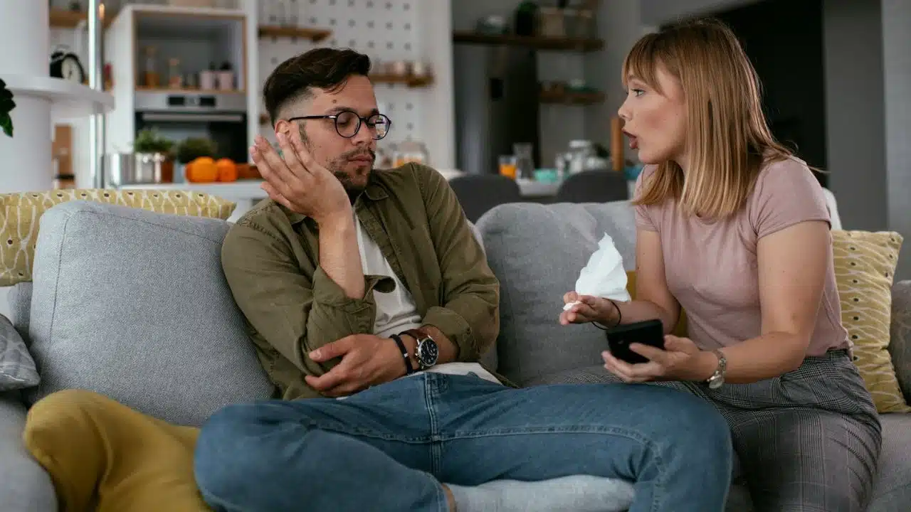 A woman in a pink shirt holds a tissue and a phone, speaking to a man in a green shirt and glasses who is sitting on a couch.