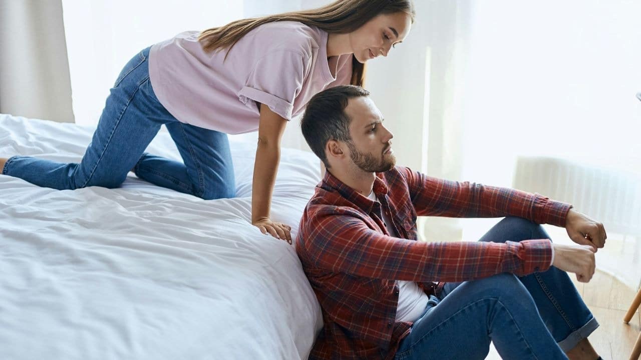 A woman in a light purple t-shirt and jeans kneels on a bed while looking down at a man in a red and black plaid shirt sitting on the floor.