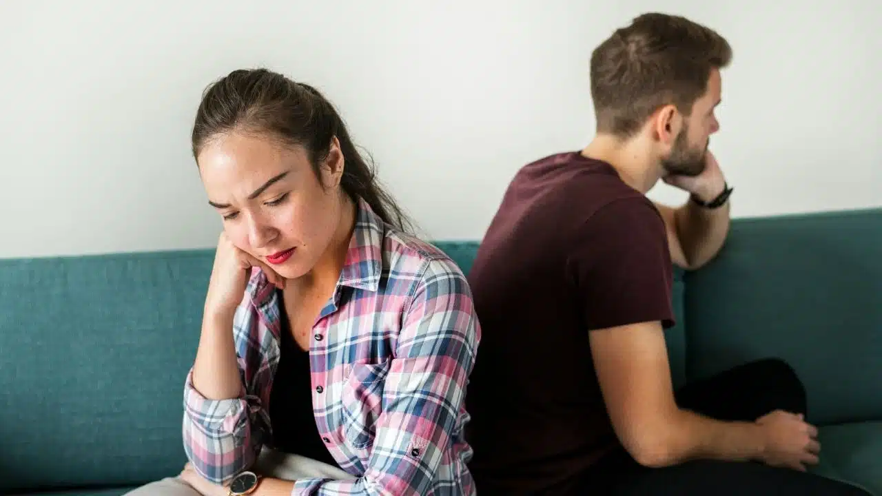 A woman sits in the foreground, looking sad, while a man faces away from her on the couch.