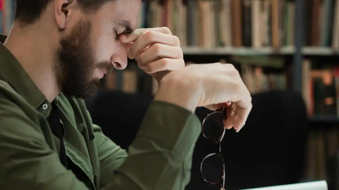 A man sitting at a desk rubbing his eyes while holding glasses.