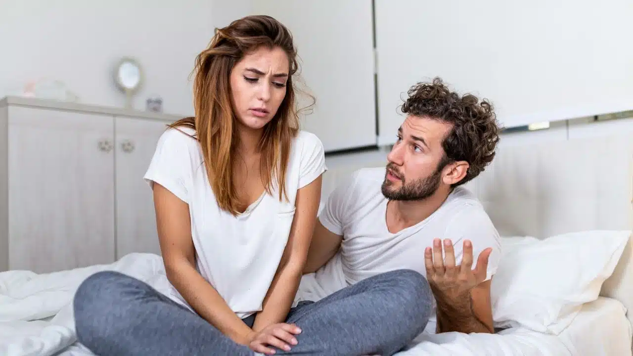 A woman in white shirt and grey pants looking distressed while a man talks to her in bed.