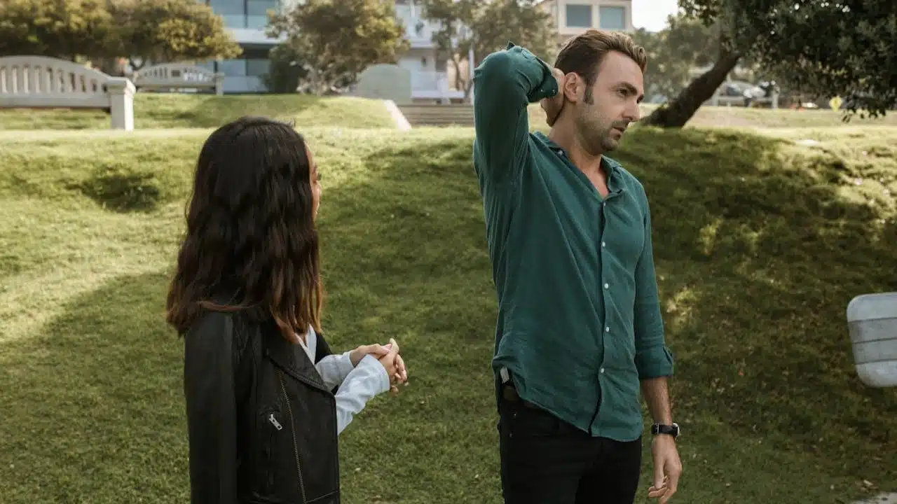 A man and woman stand on a grassy hill, and the man has his hand on the back of his neck, looking away