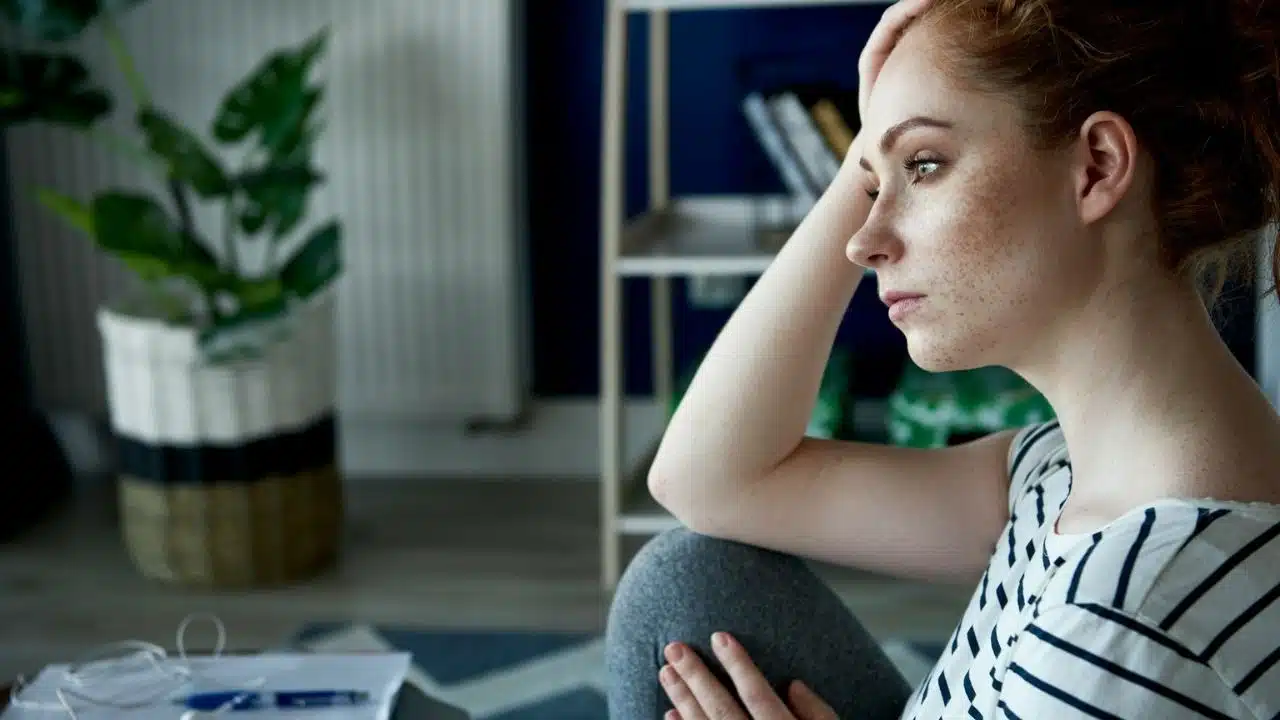 A pensive woman with freckles and red hair rests her head on her hand.