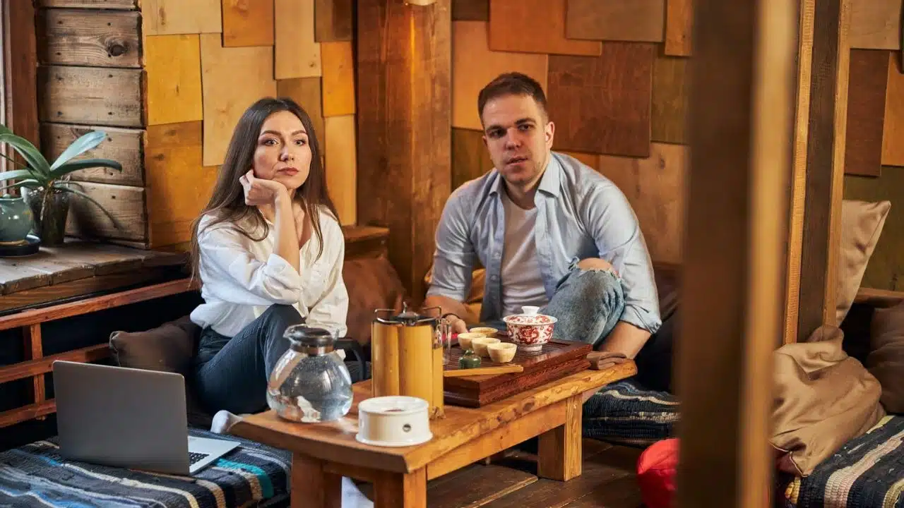 A serious man and a woman sitting on the floor are looking toward the camera.