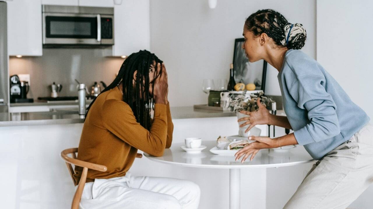 A woman gestures with her hands while a man sits at a table covering his face.