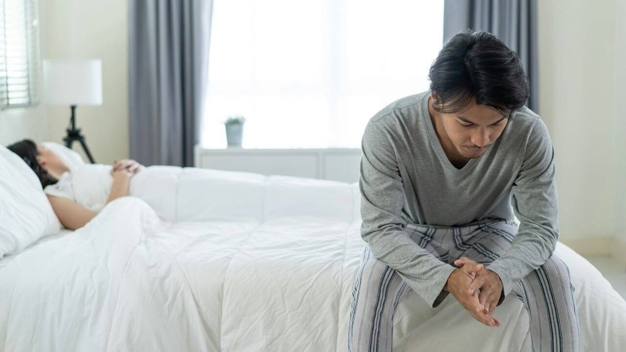 A man sits on the edge of a bed looking down while a person lies behind.