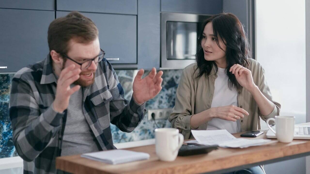A man with glasses gestures toward papers on a table while a woman looks at him.