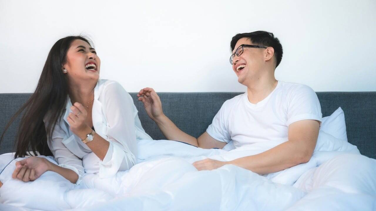 A man and woman sit in bed together, both laughing with their mouths open wide.