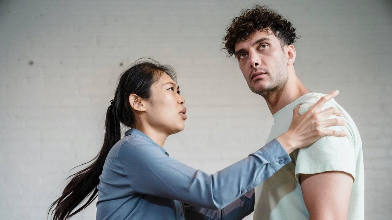 A woman with a ponytail touches a man's shoulder while speaking with a concerned expression.