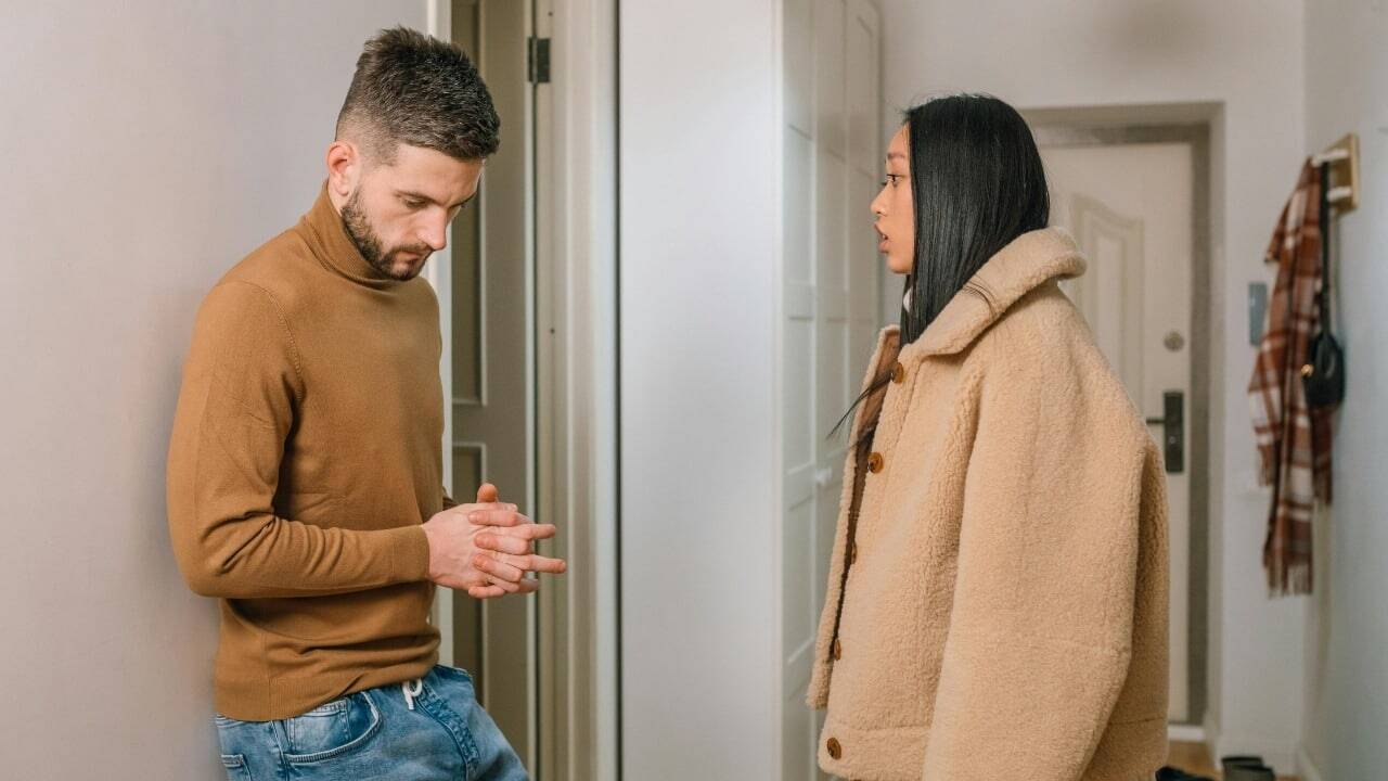 A man in a brown turtleneck looks down while a woman in a coat watches him.