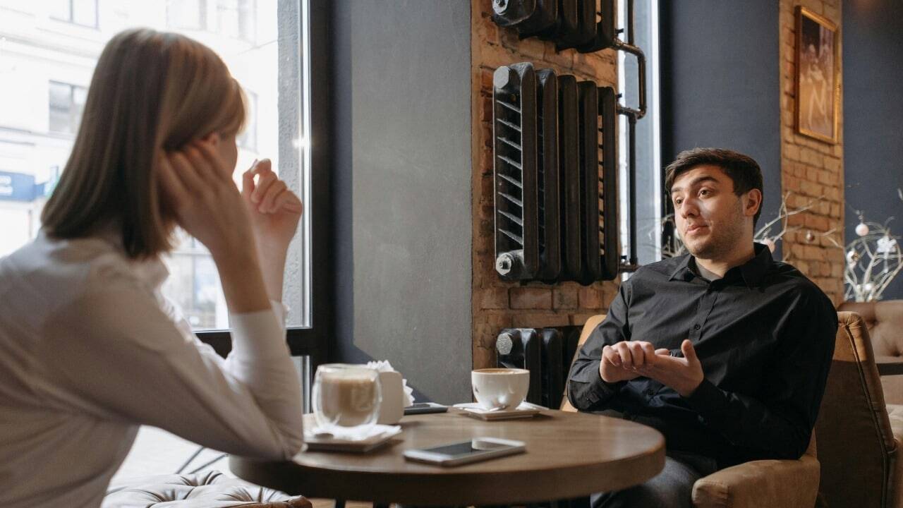 A man in a black shirt gestures with his hands while sitting across from someone.