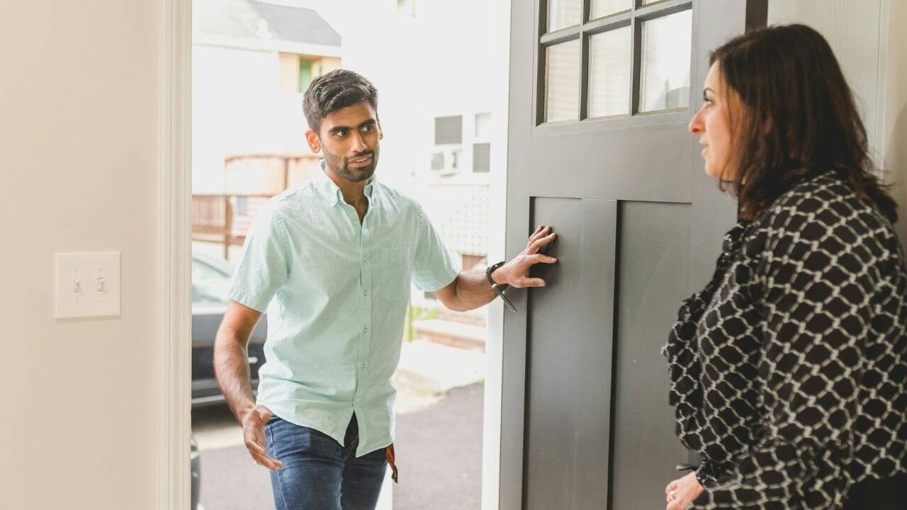 A man standing in an open doorway talks to a woman standing inside the house.