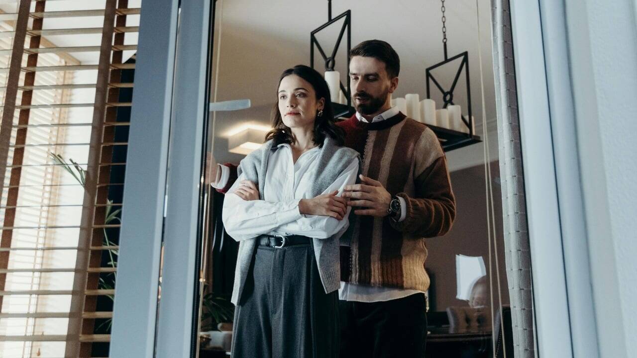 A woman stands with arms crossed while a man behind her places a hand on her arm.