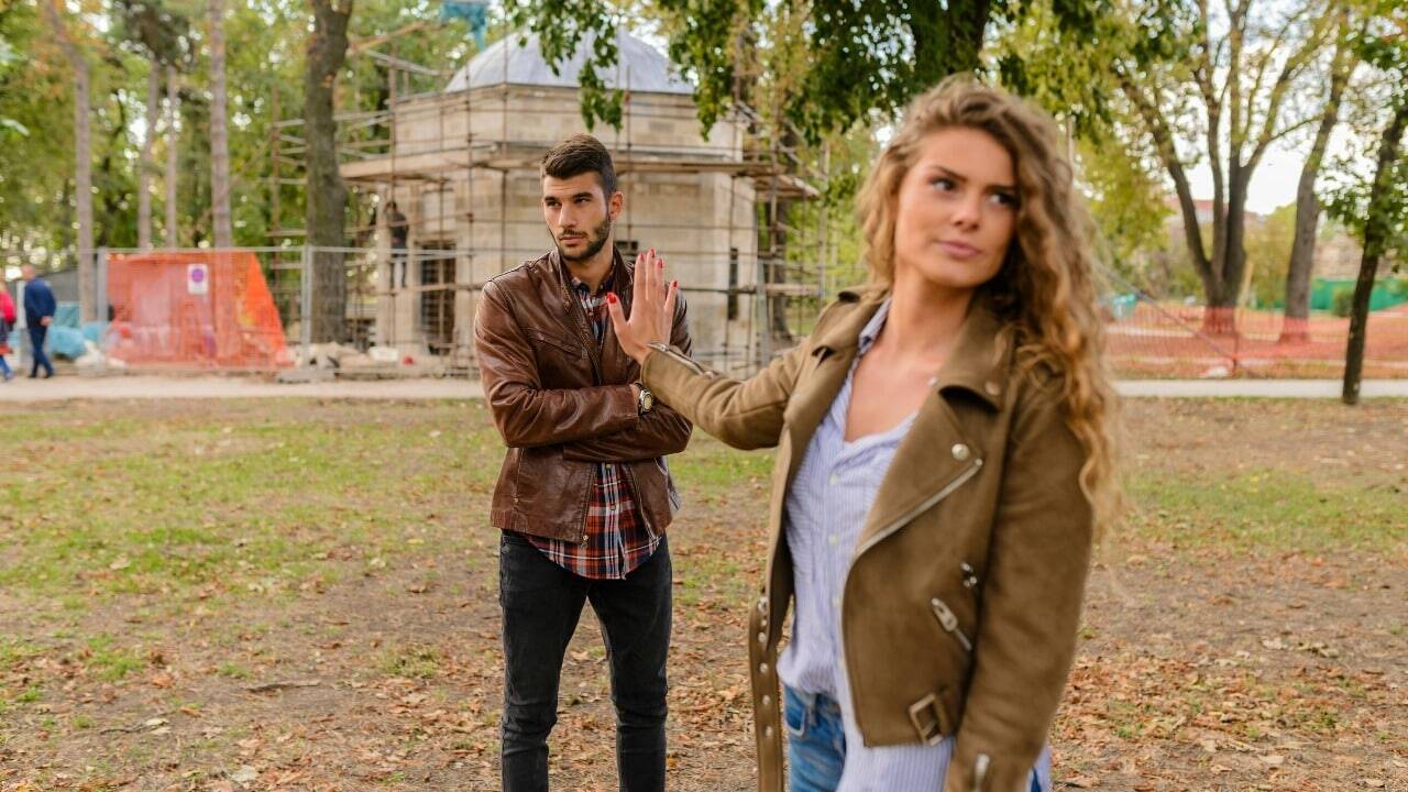 A woman holds her hand up toward a man who has his arms crossed in a park.