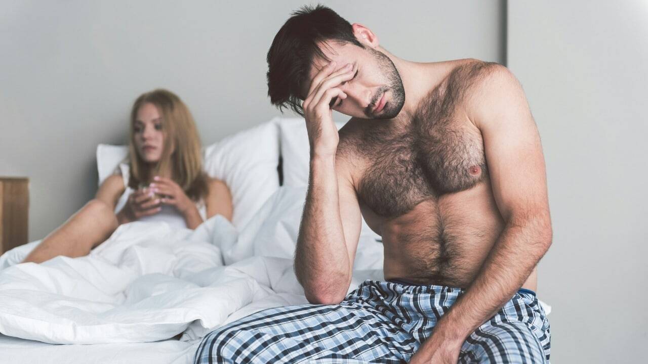 A shirtless man sits on a bed with his hand on his forehead beside someone.