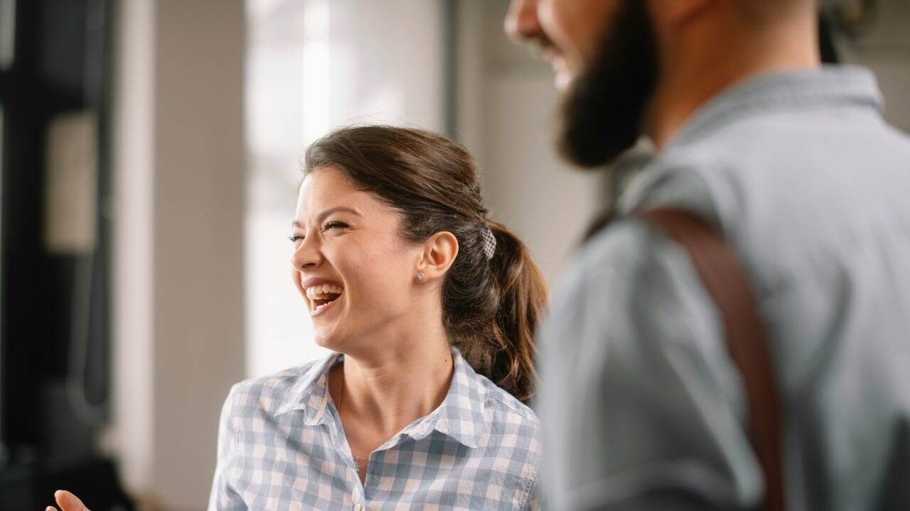 A woman with a ponytail laughs heartily while a man stands blurred in the foreground.