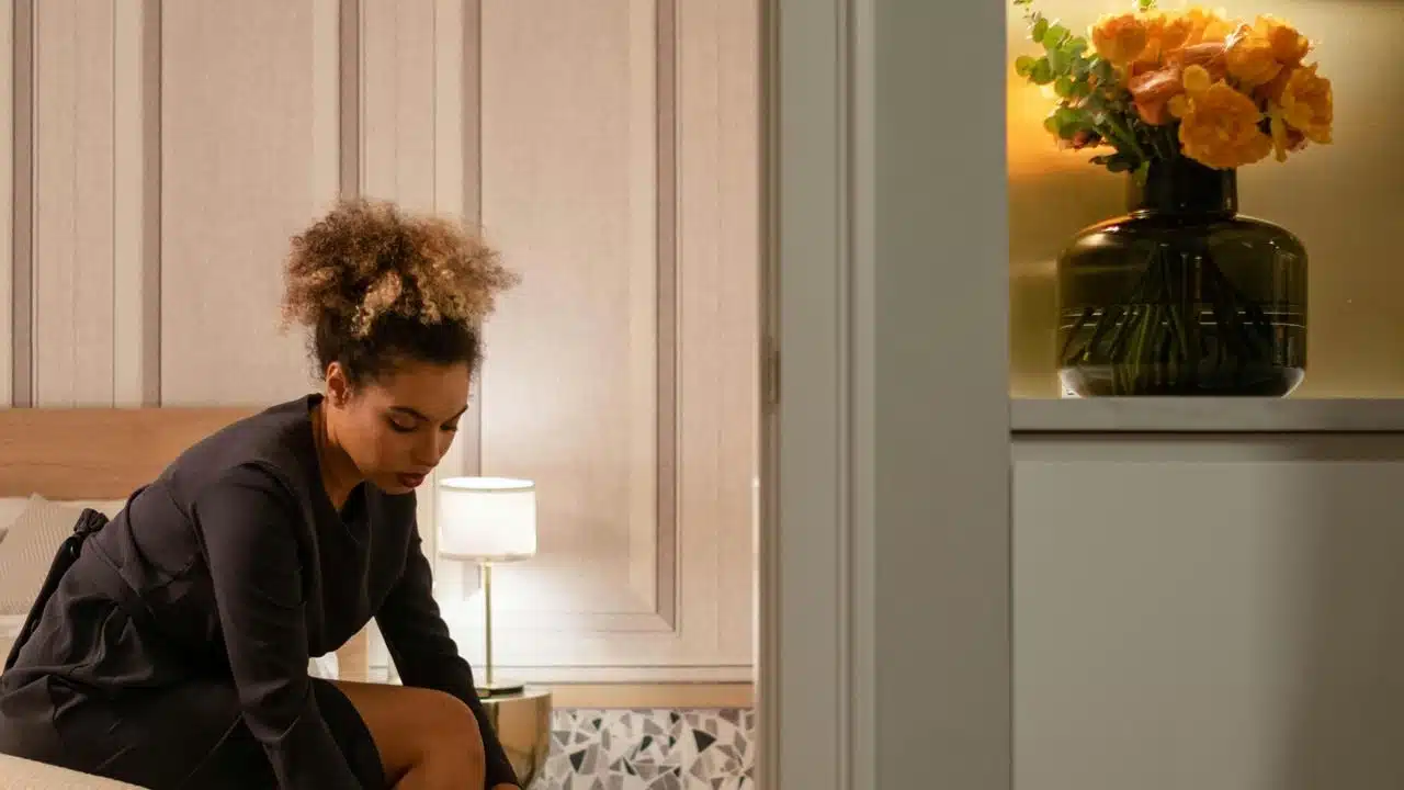 A woman sitting on a bed in a softly lit room, looking down as she adjusts something.