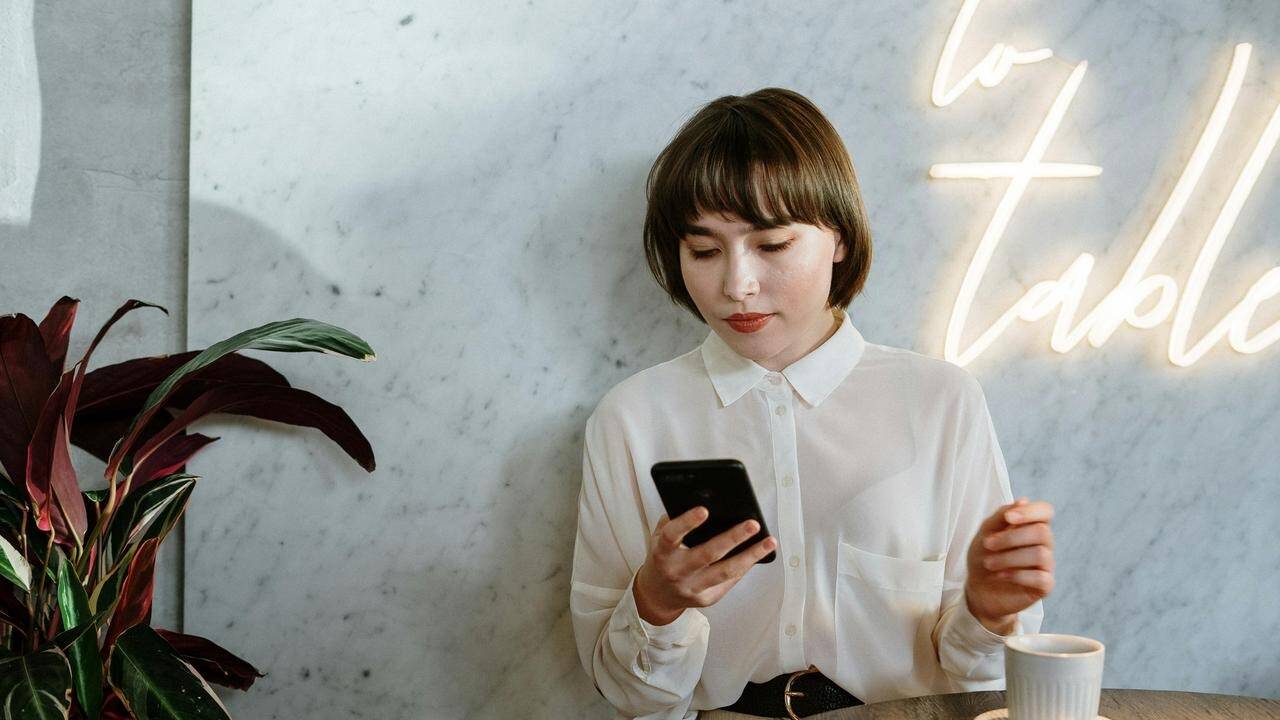 A woman sitting at a table looking at her phone with a cup of coffee nearby.