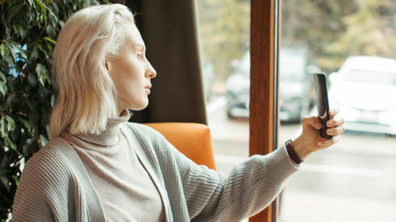 A woman with short blonde hair holding a phone near a window, looking outside.