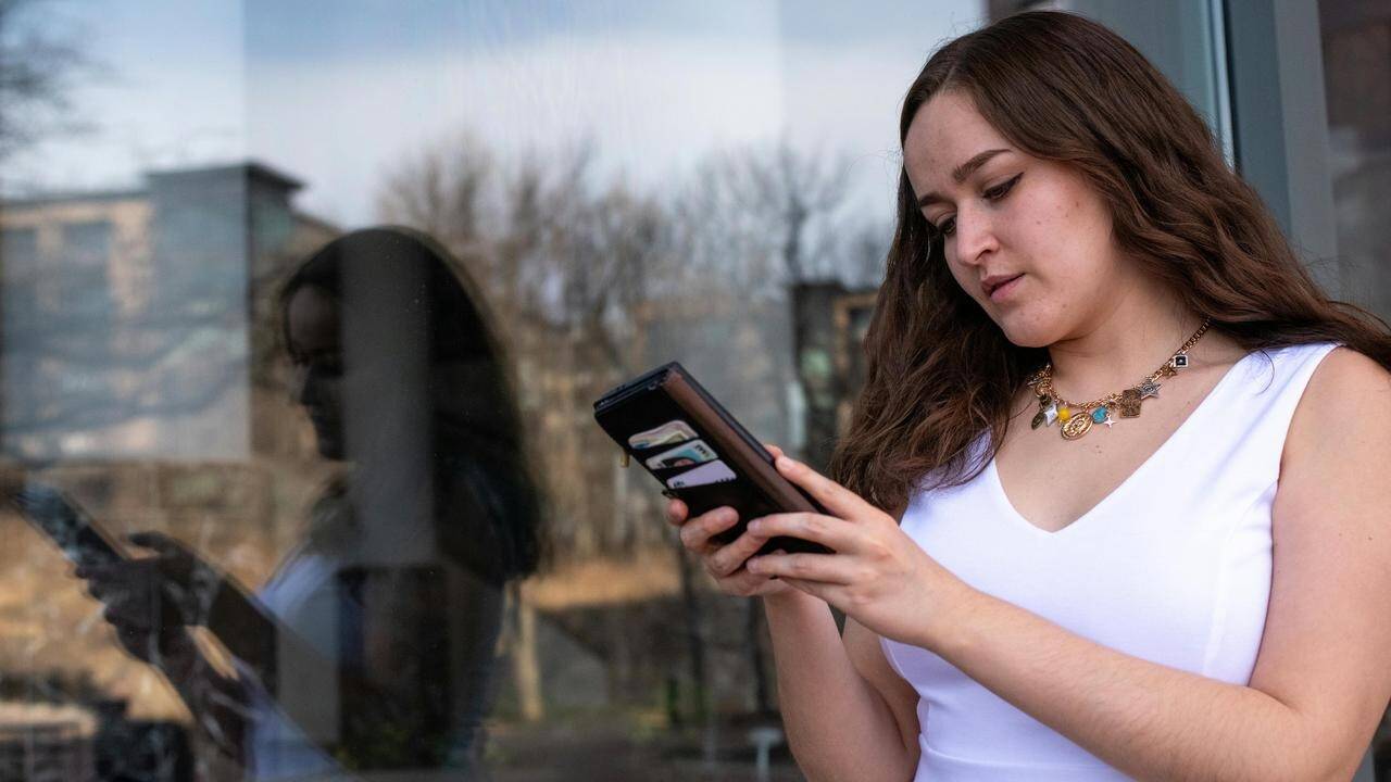 A woman standing by a window, looking at her phone with her reflection visible in the glass.
