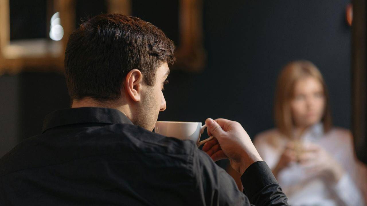 A man drinking coffee while sitting across from a woman in a dimly lit setting.