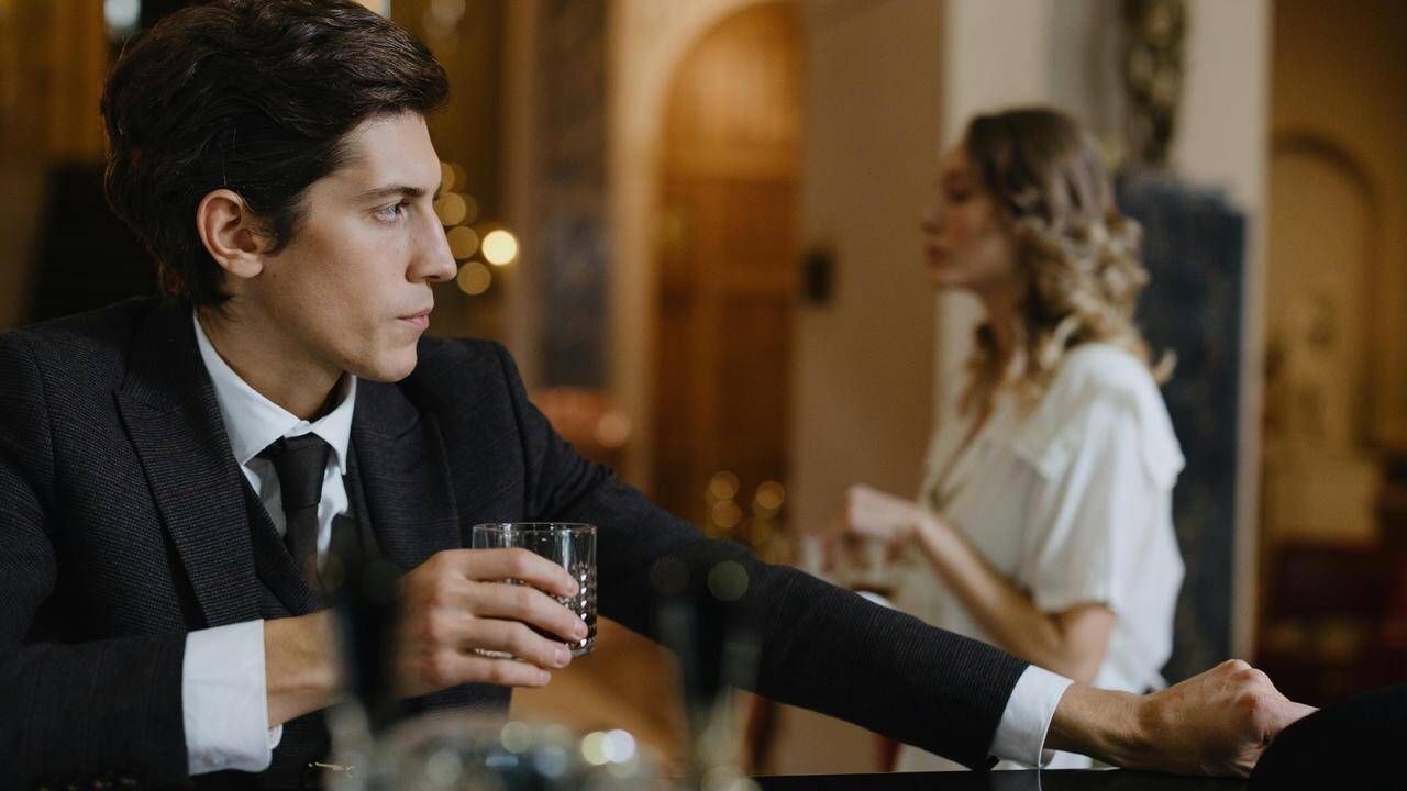 A man in a suit holding a drink at a bar while looking off to the side.