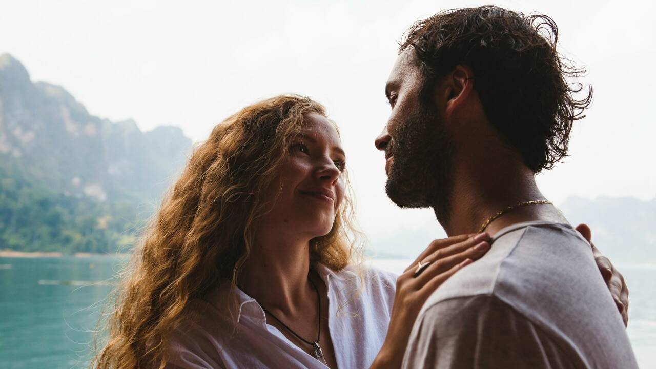 A woman with long curly hair looks up at a man standing near a lake.