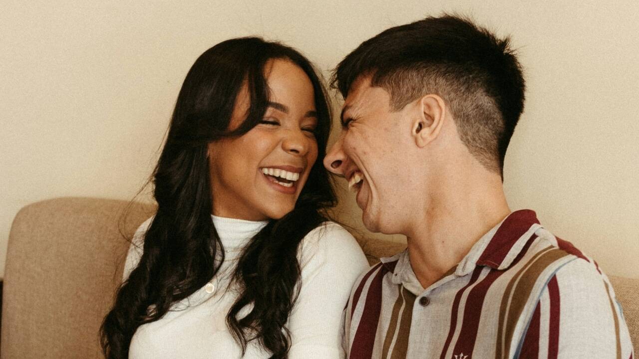 A man and a woman sit close together on a couch, laughing with their eyes closed.