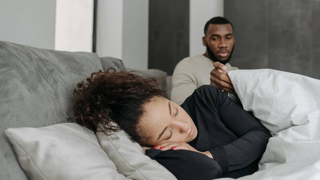 A woman sleeps on a gray sofa while a man sits behind her holding a blanket.