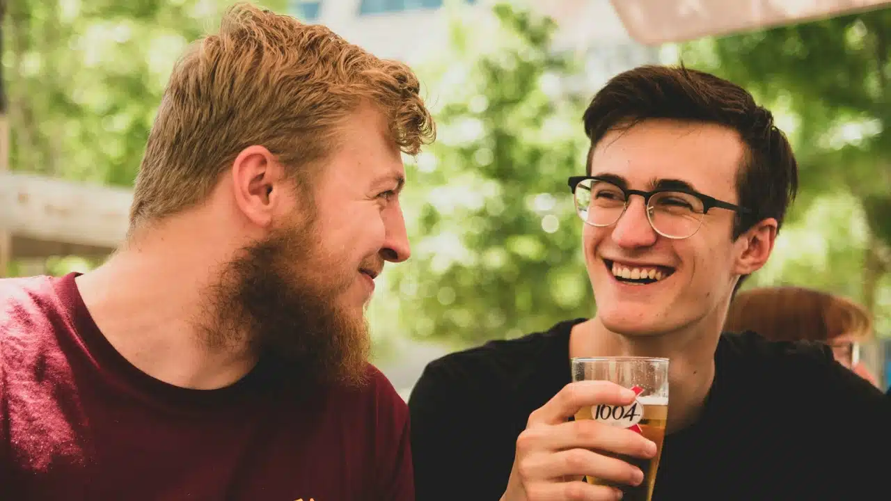 Two men sit outside, laughing together while one holds a glass containing a light beverage.