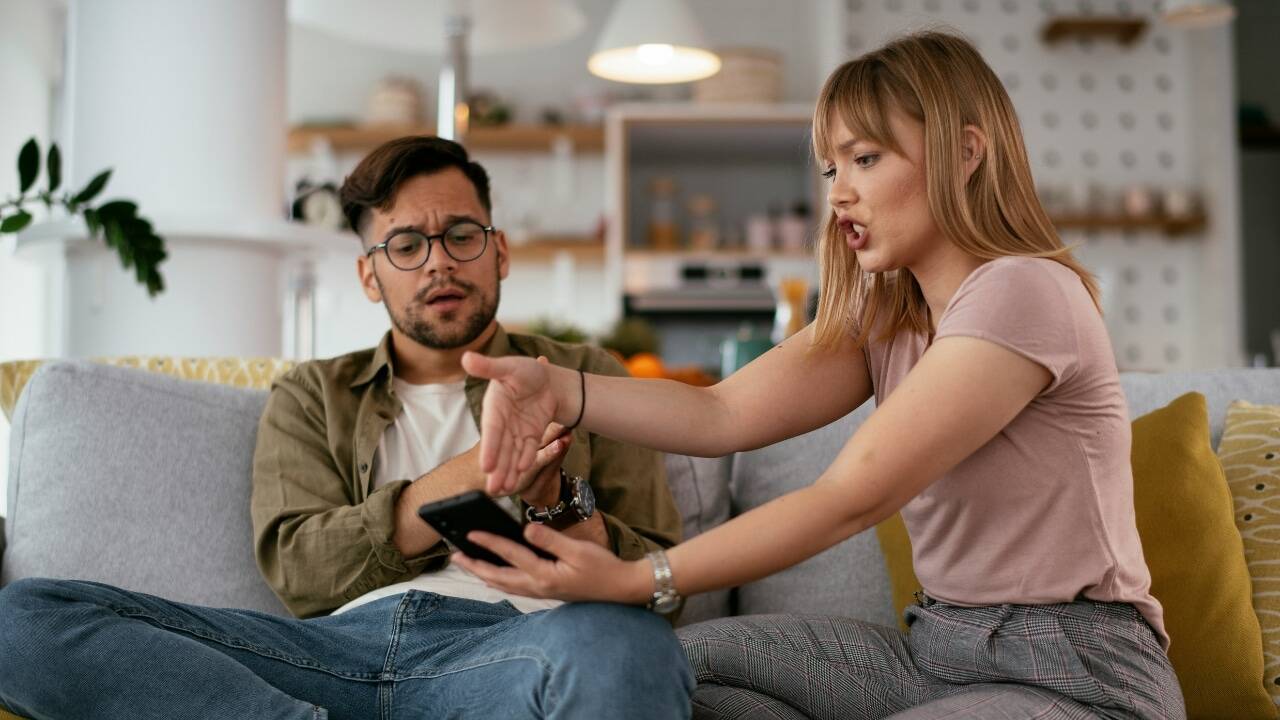 A woman on a sofa gestures toward a phone while a man looks on with concern.