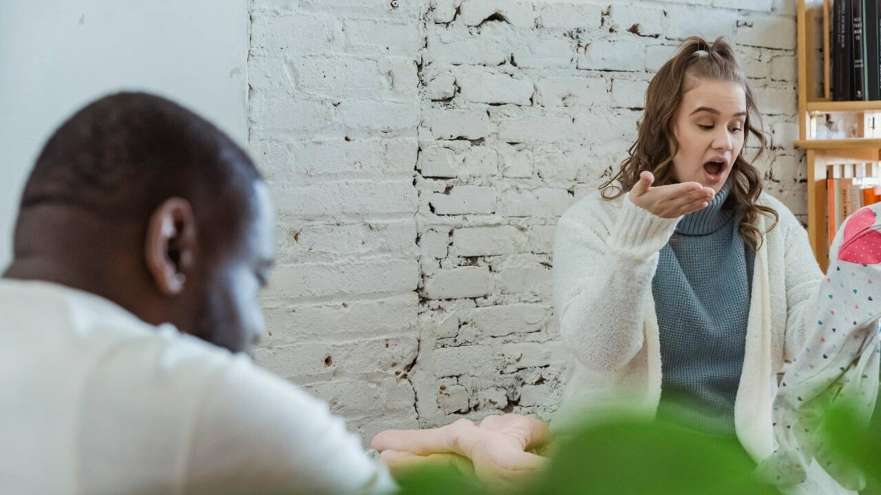 A woman in a white cardigan gestures emphatically toward a man in the foreground.