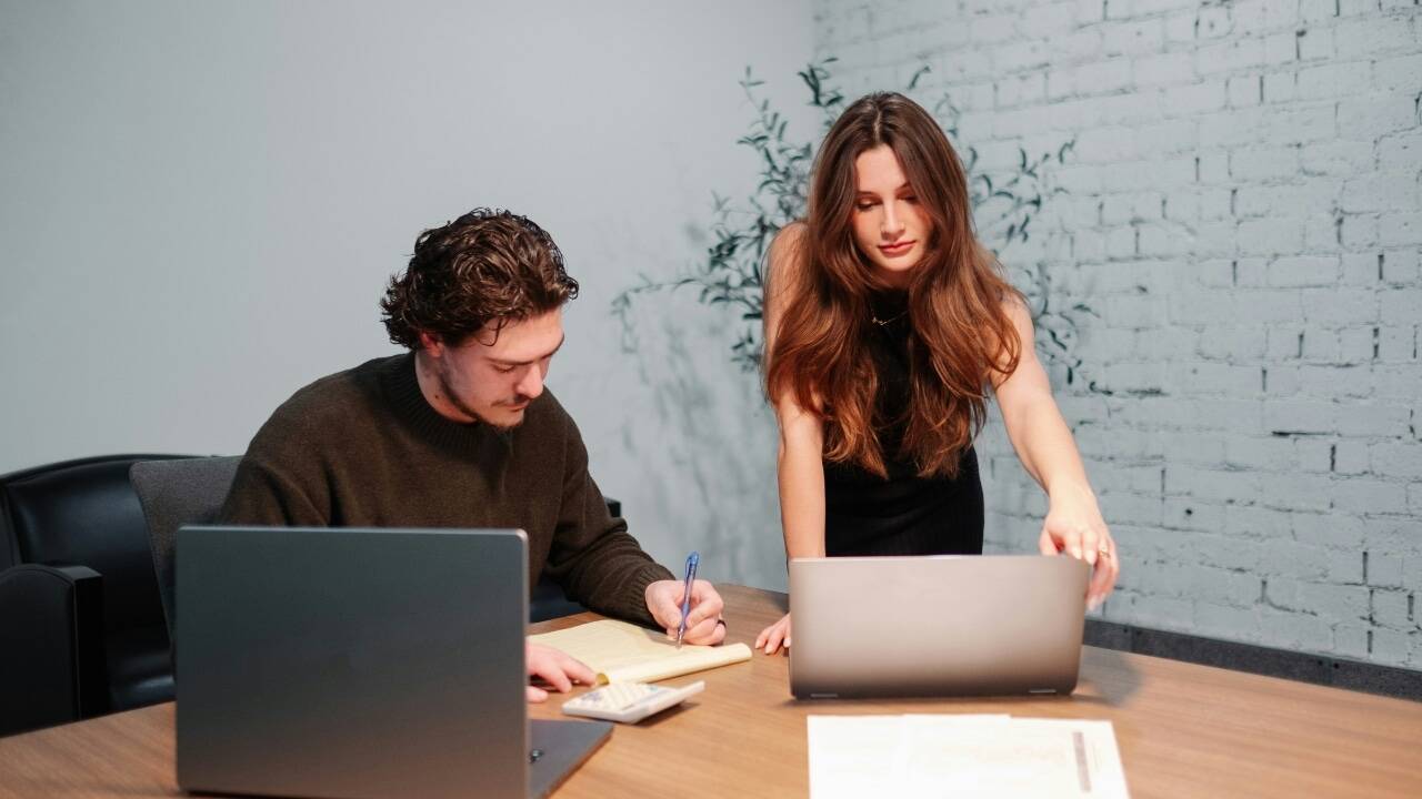 A woman stands and looks at a laptop screen while a man writes on paper.