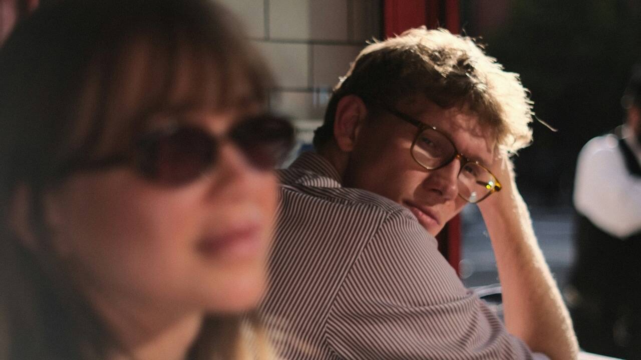 A man with glasses rests his head on his hand and looks toward a blurred woman.